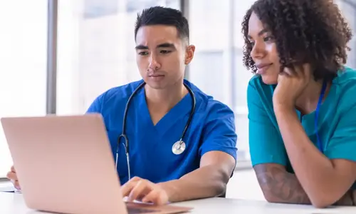 Two nurses, one male and one female, view laptop screen.