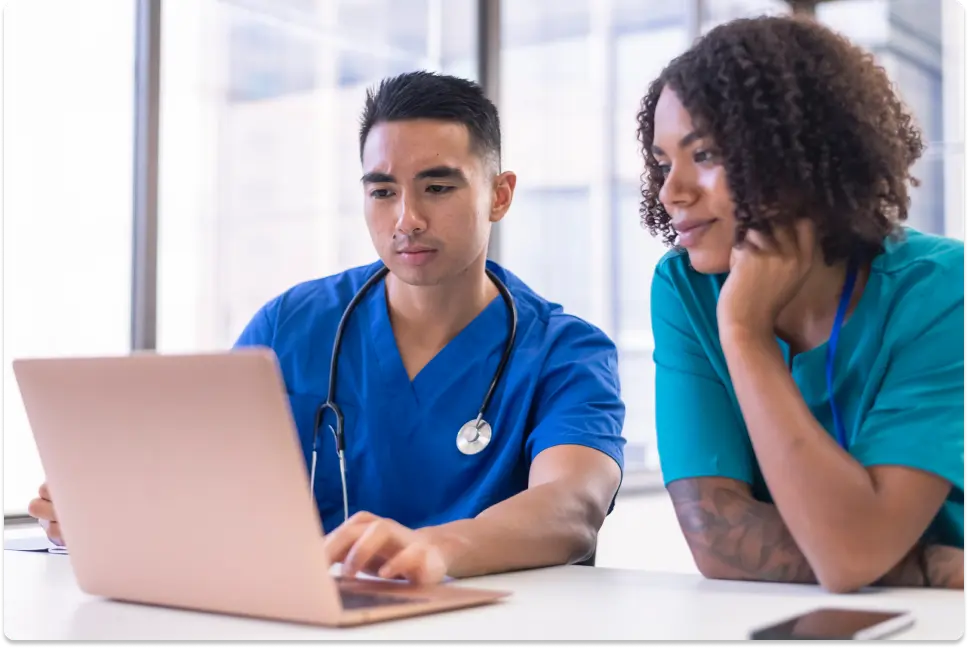 Two nurses, one male and one female, view laptop screen.