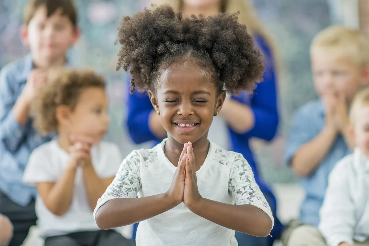 A young Black girl folds her hands in prayer in front of a group of children at a church service.