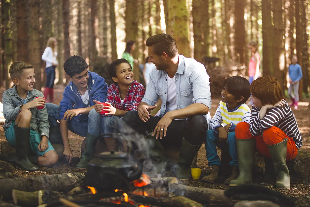 An adult volunteer overseeing a camp out sits next to a campfire with a group of smiling kids.