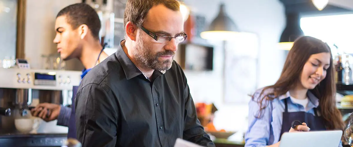 A manager in a cafe working on his tablet while teen employees work in the background.