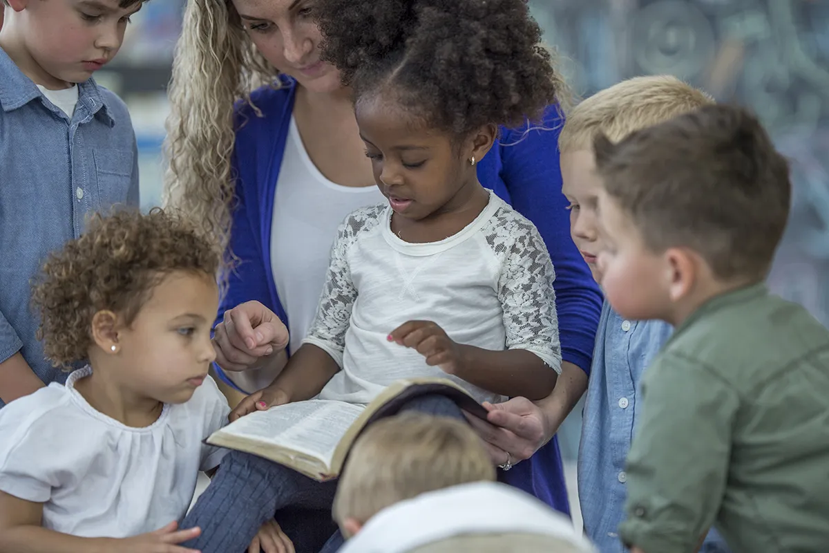 A female Sunday School teacher surrounded by children sits with a Bible on her lap.