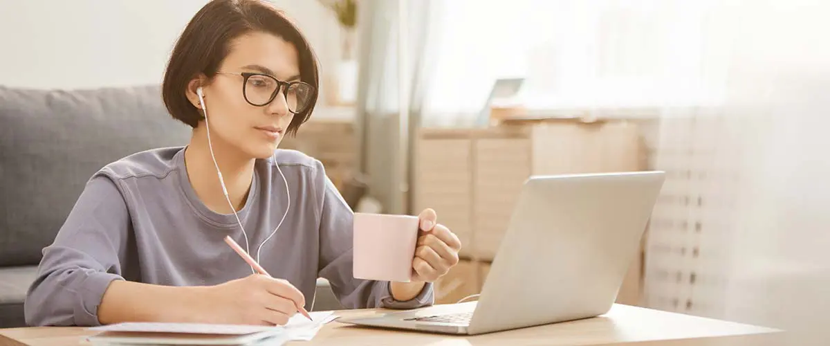 A woman taking notes at her computer while drinking a cup of coffee.