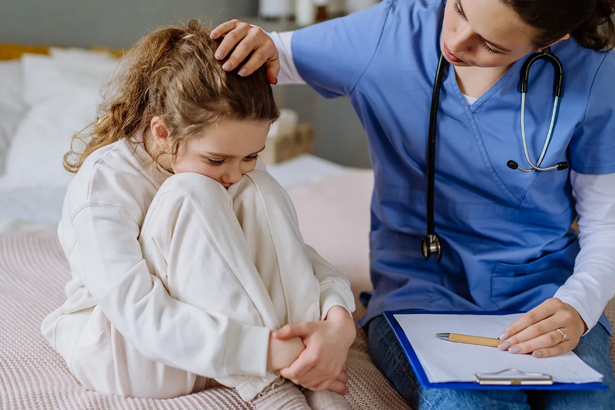 A concerned nurse wearing blue scrubs and holding a clipboard comforts a scared child.