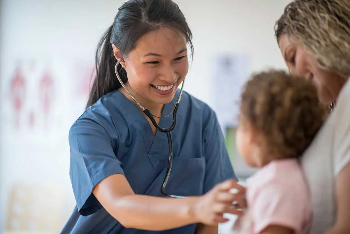 A nurse smiles as she assesses a young child. The child is sitting in their mother's lap.